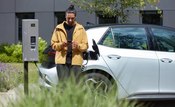 Customer stands in front of charging point and car and looks at his mobile phone